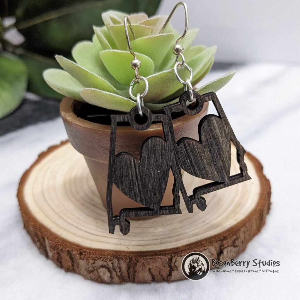 Wooden "State Line Collection" Alabama state outline earrings with heart cutouts, hanging from a small succulent potted plant posed on a slice of wood.