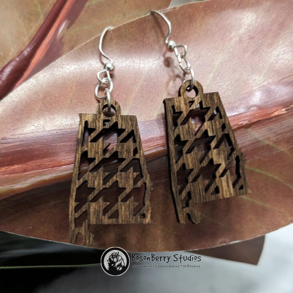 Wooden "State Line Collection" Alabama state outline earrings with houndstooth cutouts, hanging from a red magnolia leaf.