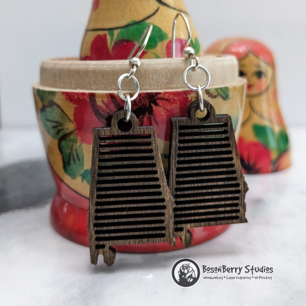Wooden "State Line Collection" Alabama state outline earrings with variegated cutouts, hanging from a wooden, yellow and red nesting doll on a marble backdrop.