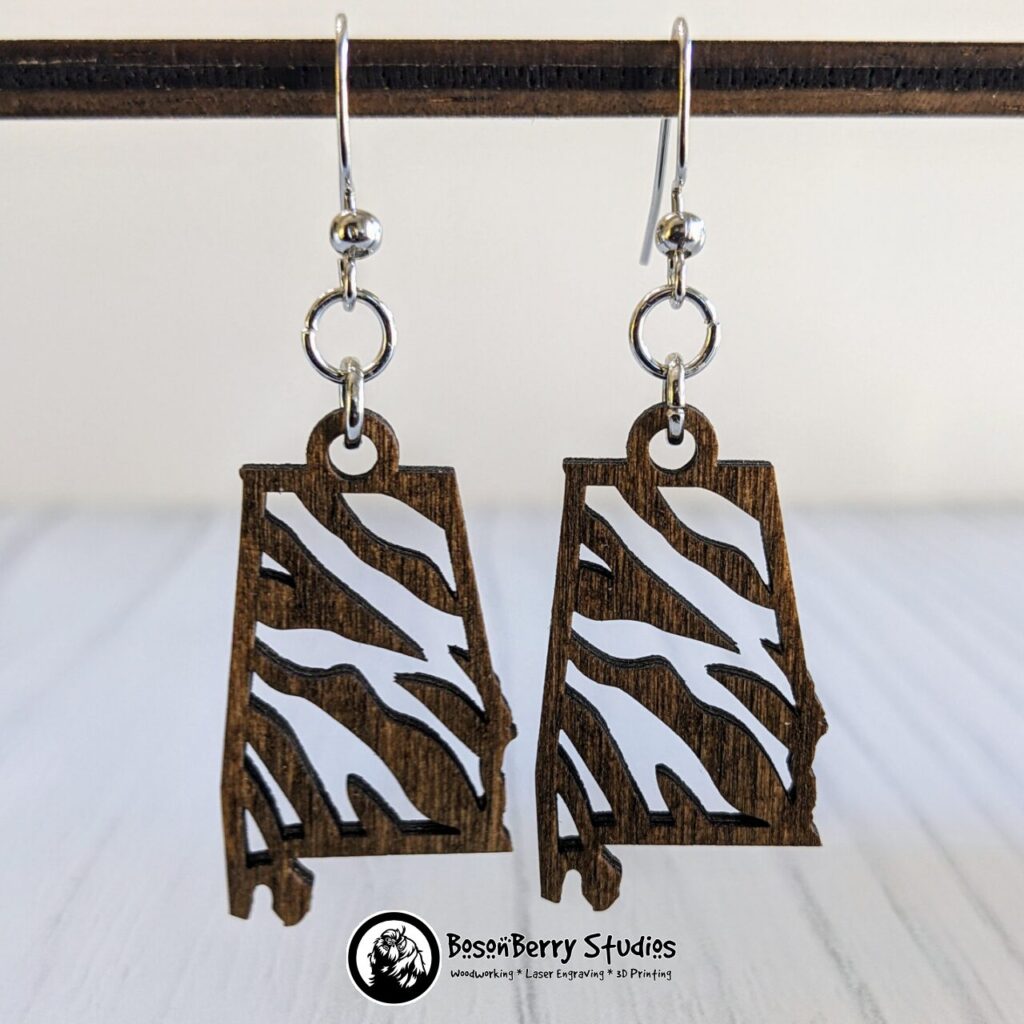 Wooden "State Line Collection" Alabama state outline earrings with tiger stripe cutouts, hanging from a wooden dowel over a white backdrop.