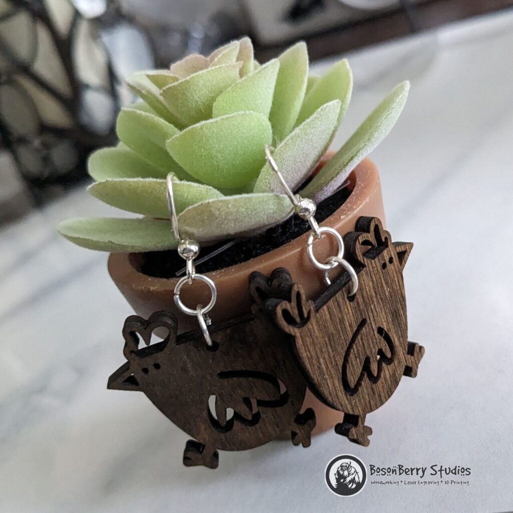 Cute Wooden cartoony Chicken earrings, facing opposite directions, posed on a tiny potted succulent plant on a marble backdrop.
