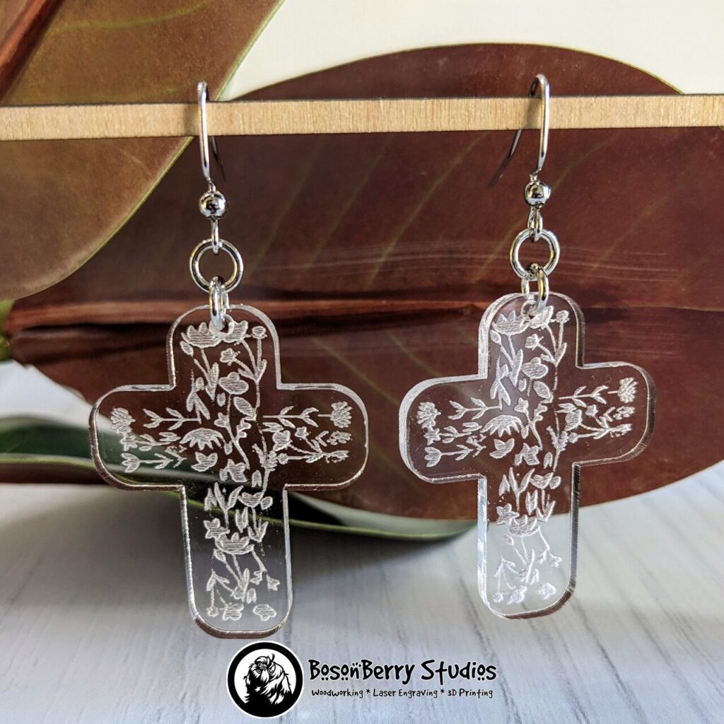 Clear Acrylic cross shaped Earrings with white floral pattern engraved on them, hanging from a wooden dowel in from of red magnolia leaves.