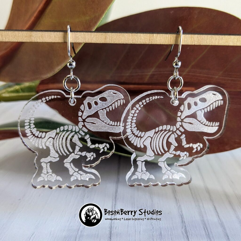 Clear Acrylic Earrings with a white T-Rex Dinosaur Skeleton engraved on them, hanging from a wooden dowel with a backdrop of red magnolia leaves.