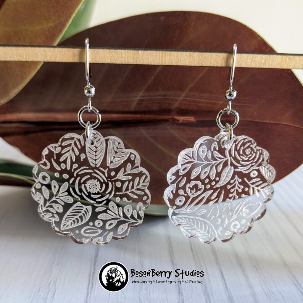 Clear Acrylic Scalloped shaped Earrings with white floral leaf pattern engraved on them, hanging from a wooden dowel in from of red magnolia leaves.