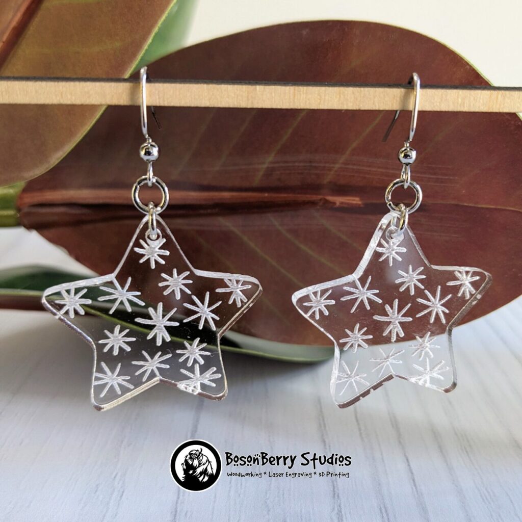 Clear Acrylic Star shaped Earrings with white asterisks engraved on them, hanging from a wooden dowel in from of red magnolia leaves.