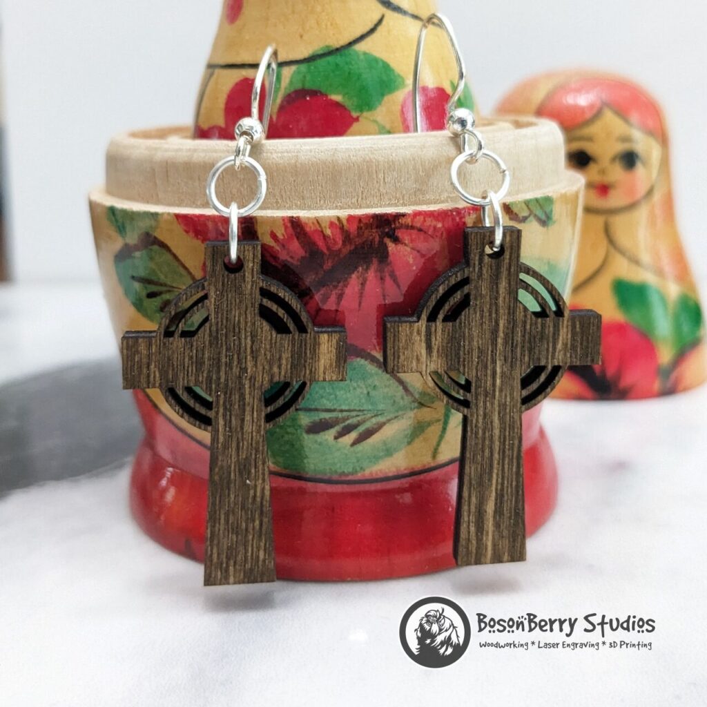 Wooden Radiant Cross Earrings, stained brown, hanging from a red yellow wooden nesting doll on marble backdrop.