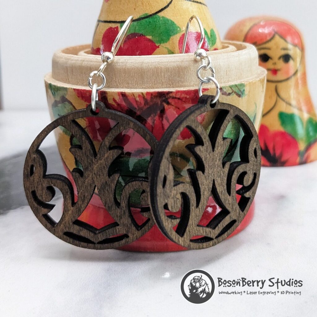 Damask Cutout Wooden Earrings, round shaped, brown stained, hanging from a red yellow wooden nesting doll on a marble backdrop.