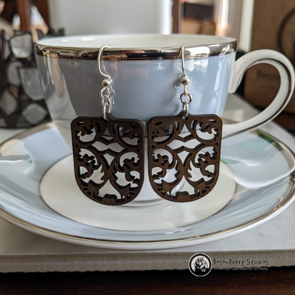 Damask Cutout Wooden Earrings, shield shaped, brown stained, hanging from a blue white and silver china teacup on a matching saucer.