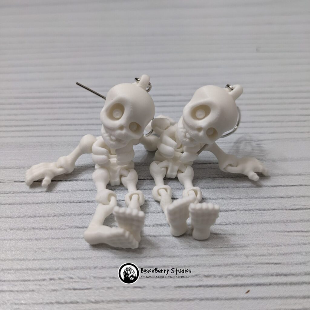 3D printed flexible cartoon Skeleton earrings made of white filament posed on a white wood plank.