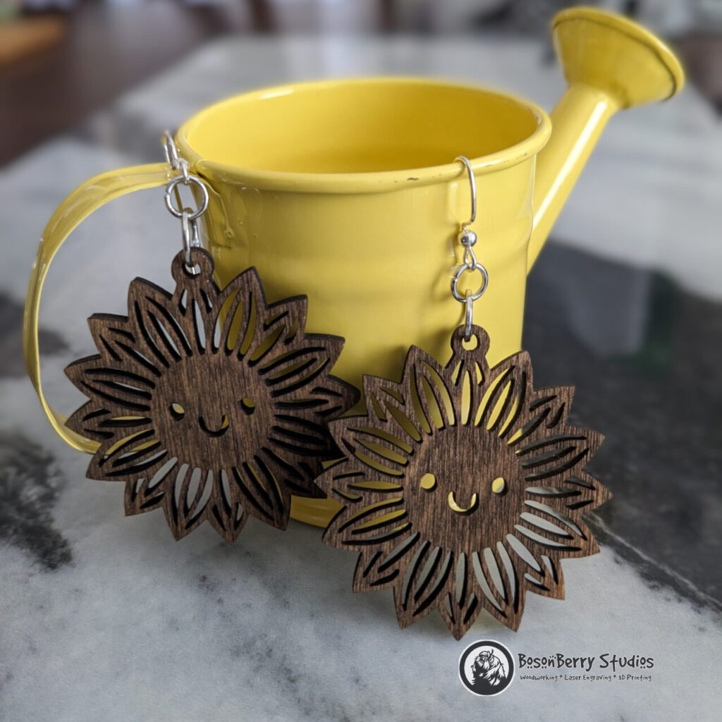 Wooden Sunflower Earrings with smiley face cutouts in the center, stained brown, hanging from a tiny, yellow metal watering can on a marble backdrop.