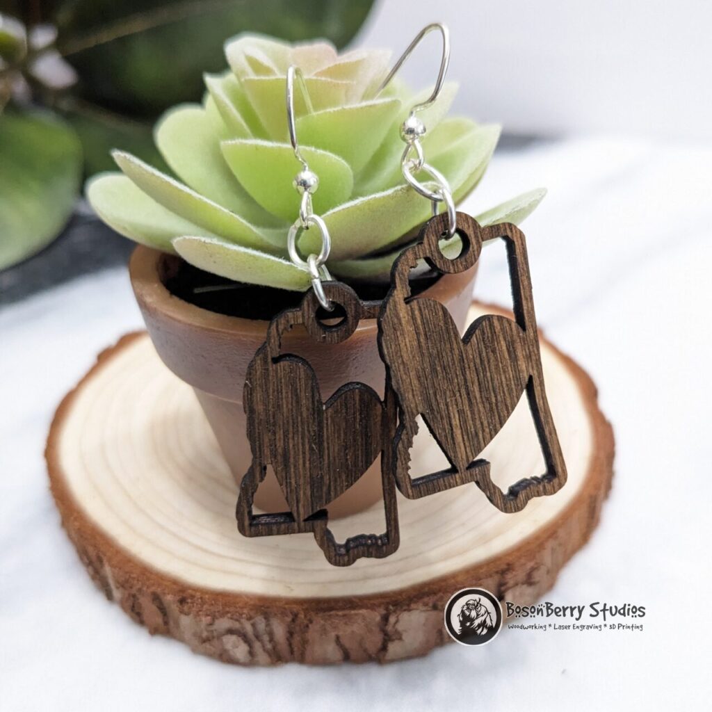 Wooden "State Line Collection" Mississippi state outline earrings with a heart cutout filling the center, hanging off a miniature succulent potted plant posed on a slice of wood.