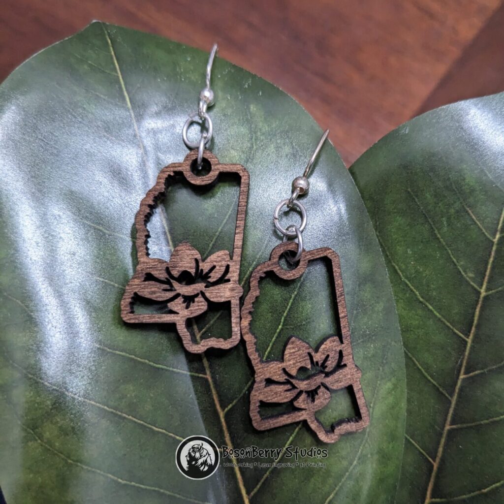 Wooden "State Line Collection" Mississippi state outline earrings with a magnolia flower cutout in the middle, hanging from magnolia leaves on a wooden background.