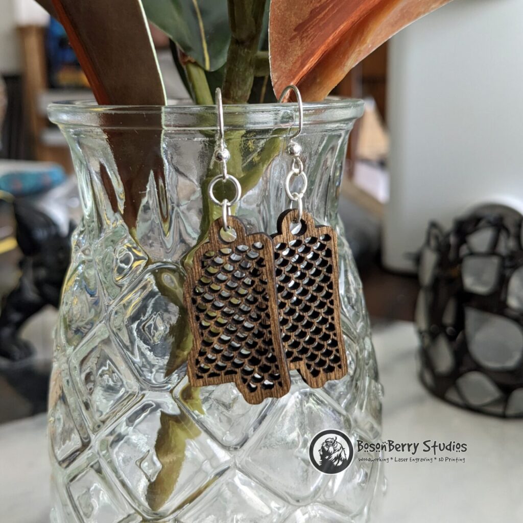 Wooden "State Line Collection" Mississippi state outline earrings with fish scale cutouts, hanging off a clear, decorative pineapple glass accented by magnolia leaves.