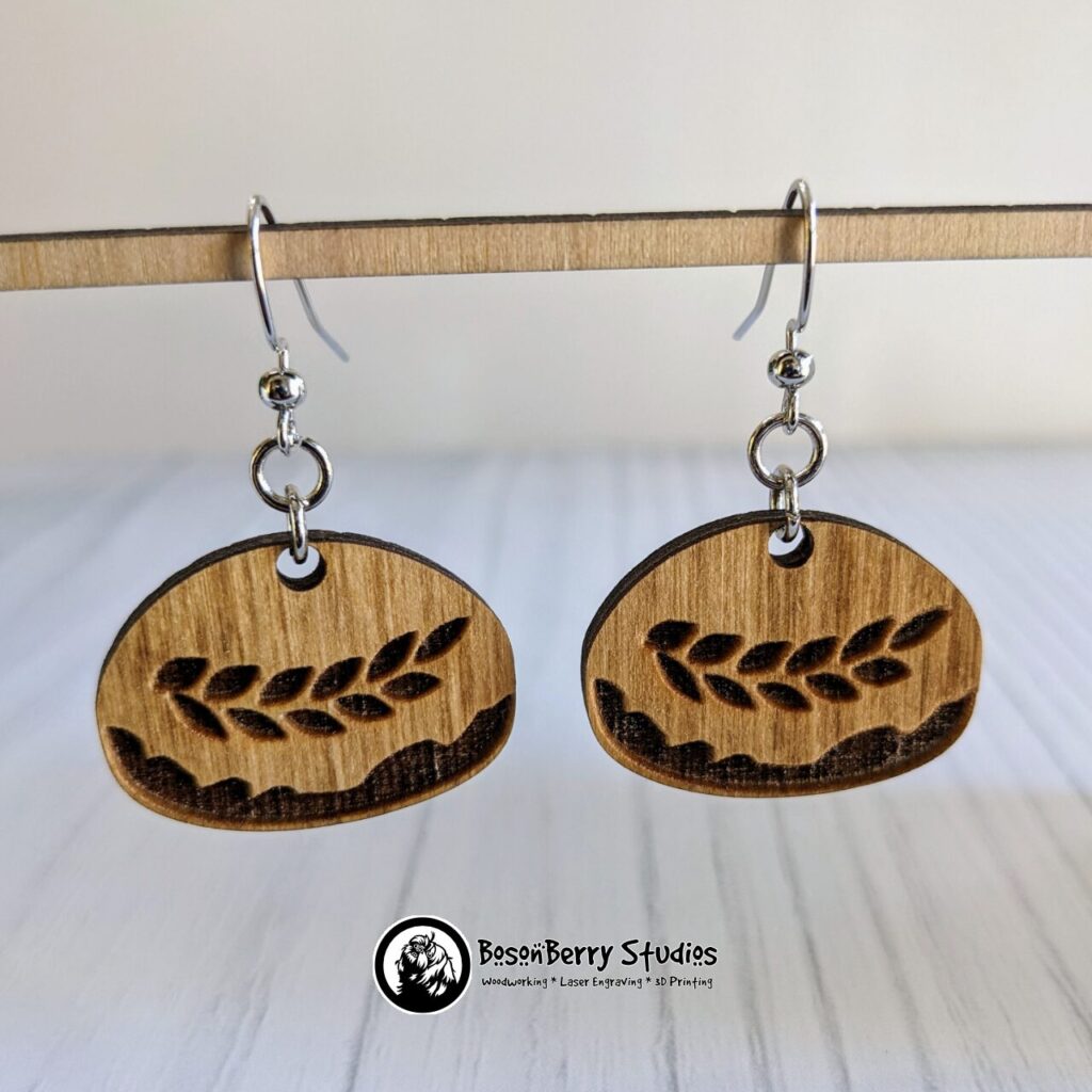 Wooden Sourdough loaf shaped earrings with horizontal laurel pattern hanging from wooden dowel in front of a white backdrop.