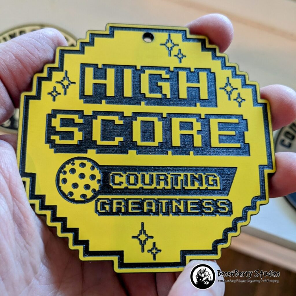 High Score: Courting Greatness acrylic bag tag. Pixelated yellow acrylic coin with black engraves featuring the text High Score: Courting Greatness, a pickleball, and pixelized sparkles. Shown held in an adult's hand.