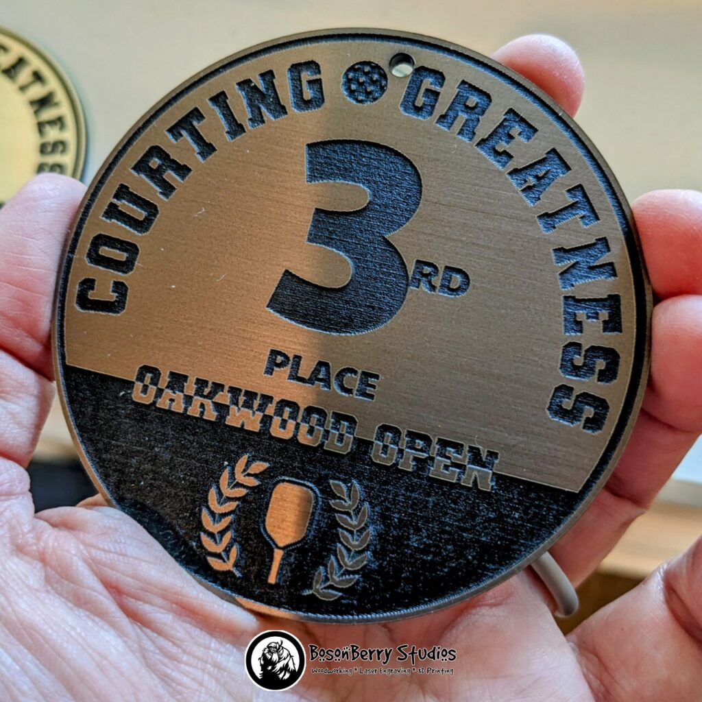 Bronze/Black Acrylic Bag Tag Keychain. Circular and features the words "Courting Greatness 3rd Place Oakwood Open" with a pickleball, a paddle, and a laurel wreath. Shown held in an adult hand.