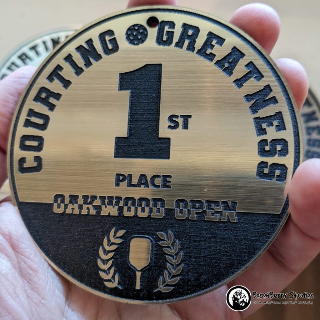 Gold/Black Acrylic Bag Tag Keychain. Circular and features the words "Courting Greatness 1st Place Oakwood Open" with a pickleball, a paddle, and a laurel wreath. Shown held in an adult hand.