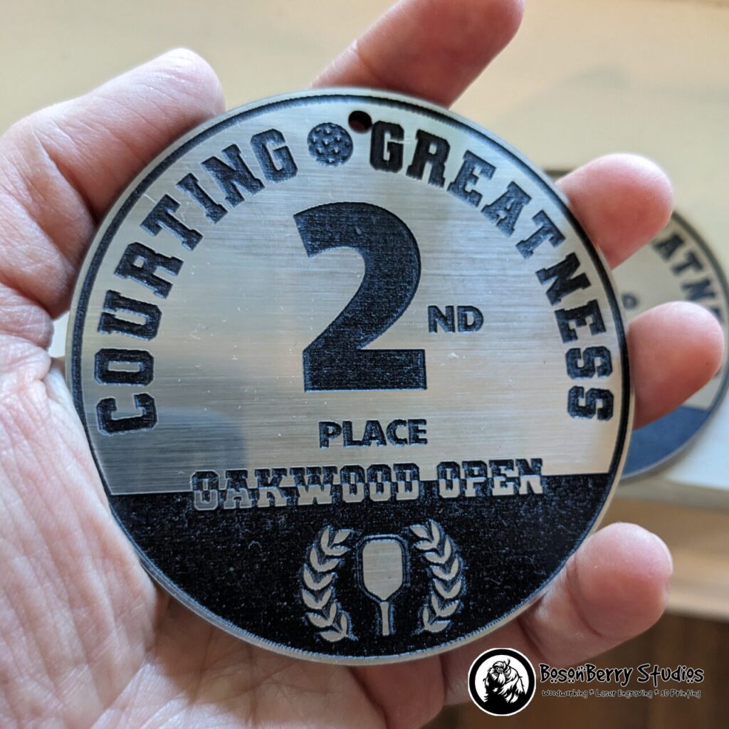Silver/Black Acrylic Bag Tag Keychain. Circular and features the words "Courting Greatness 2nd Place Oakwood Open" with a pickleball, a paddle, and a laurel wreath. Shown held in an adult hand.
