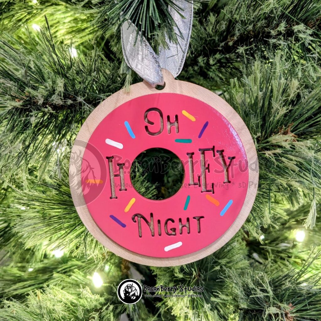 "Oh Holey Night" Doughnut Premium Wood Christmas Ornament. Layered with round wooden backer with hole in center. Front layer is round doughnut painted pepto pink, with words "Oh Holey Night" cut out. The O in Holey is the center hole of the donut. Sprinkles in blue, purple, white, orange and yellow and hand-painted all over the doughnut. Designed and made by BosonBerry Studios.