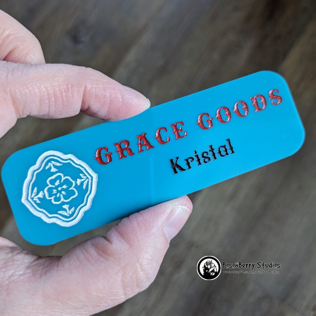 Rectangular, paint-filled turquoise acrylic name tag. A floral logo on the left is filled with white paint. The name Grace Goods is filled with red. The name Kristal is filled with black. A hand holds the name tag over a wooden backdrop.
