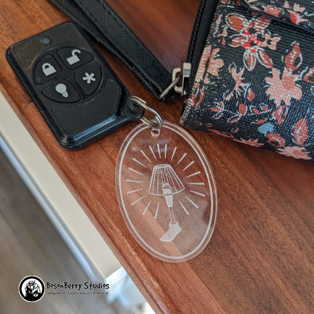 Clear acrylic oval keychain engraved with an artificial leg with a lampshade on it, and light beams radiating from it. Displayed with a keyfob remote and a floral wallet on a wooden countertop.
