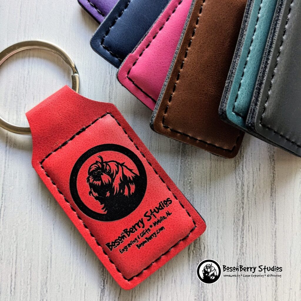 A red leatherette keychain with BosonBerry Studios logo and info engraved on it in black, flanked by various other colors of leatherette keychains, on a white wood backdrop.