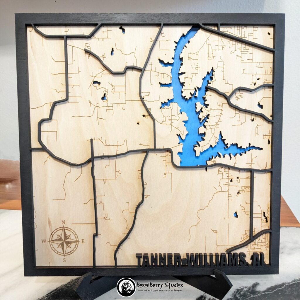 Map of Tanner Williams, AL made of laser-cut wood. Back blue layer shows Big Creek Lake, center layer is birch colored with minor roads and compass road engraved, top layer is black border, with major highways and title "Tanner-Williams, AL" in bottom right corner.