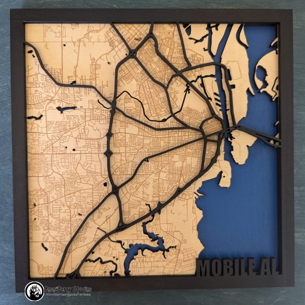 Map of Mobile, AL made of laser-cut wood. Back layer for bay/river water is blue, middle layer is birch colored with minor roads engraved, top layer is black border with black major highways, and title "Mobile AL" in bottom right corner.