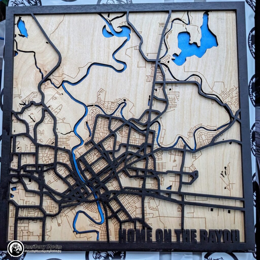 Map of Monroe, LA, made of laser-cut wood. Back lake/river layer is blue, middle is birch colored, engraved with minor roads, top is black border with black major highways and title "Home on the Bayou" in bottom right corner.