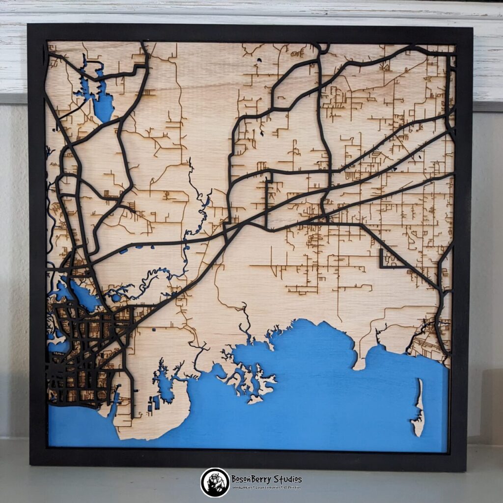 Map of Pascagoula, MS, made of laser cut wood. Back layer is blue for water, middle layer is birch colored and engraved with brown minor roads, top layer is a black border with black major highways.