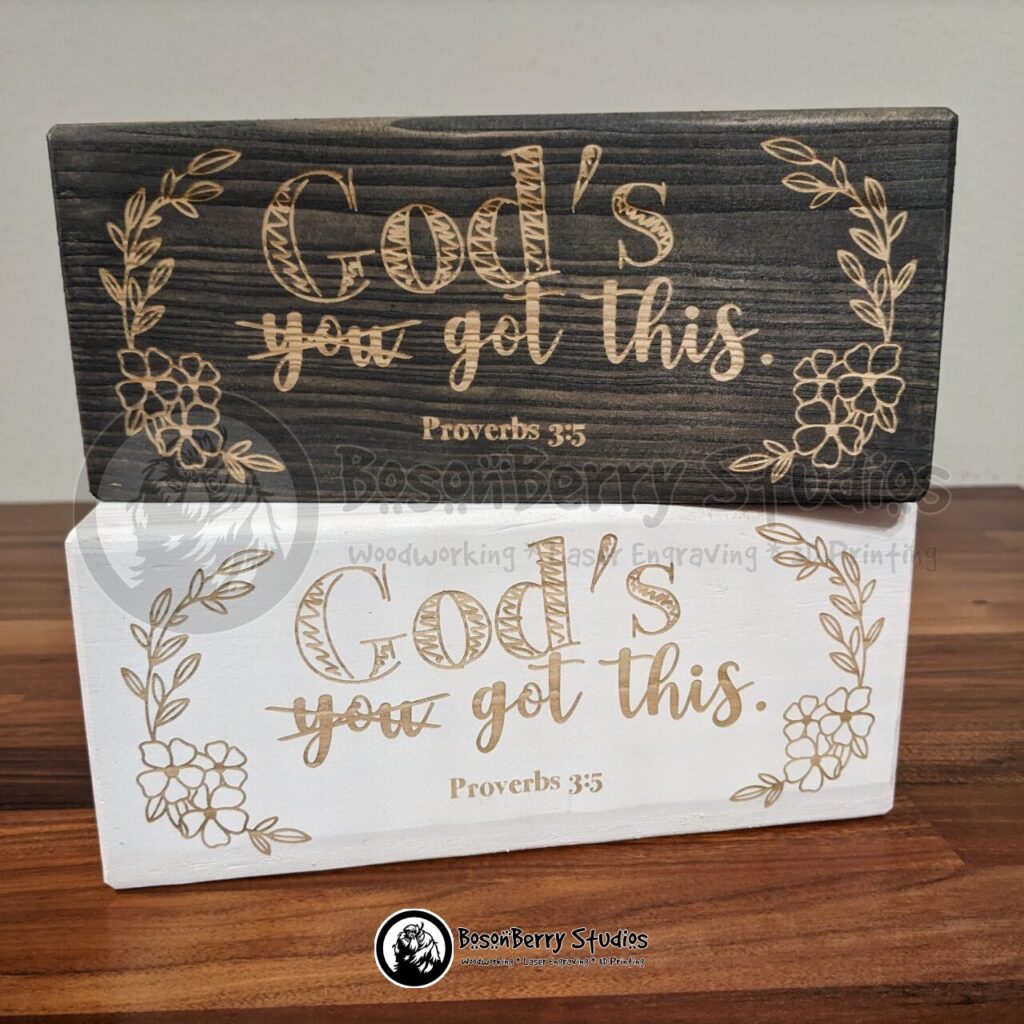 Bible Blocks by BosonBerry: Proverbs 3:5. Large, horizontal wooden blocks shown in both grey stain and white paint versions, engraved with text "you got this", only the word "you" is crossed out and the word "God's" written in sketchy bold letters above it; floral illustrations are on either side of the text. Created by BosonBerry Studios.
