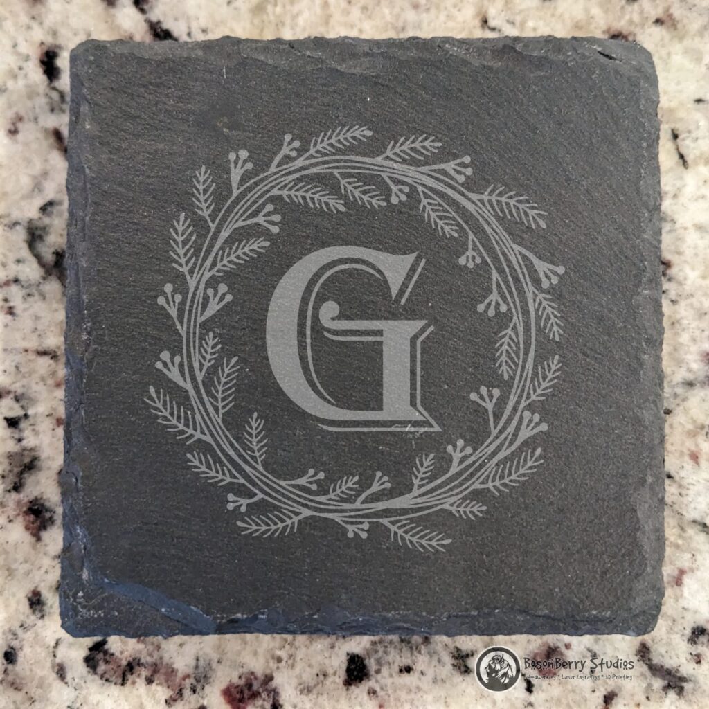 Herb Wreath Monogram Slate Coasters. An ornate letter G with a shadow is in the center of a wreath of herbs and tiny berries, in the middle of a square, gray, slate coaster. Displayed on a quartz backdrop.