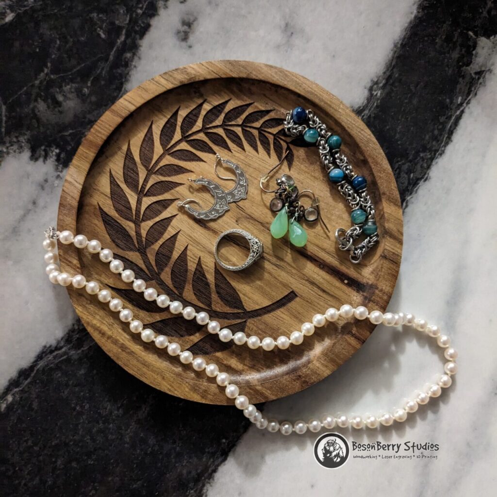Small Round Acacia Jewelry Tray engraved with round laurel leaf design by BosonBerry Studios. Tray is scattered with earrings, a ring, a bracelet and a pearl necklace. Black and white marbled backdrop.