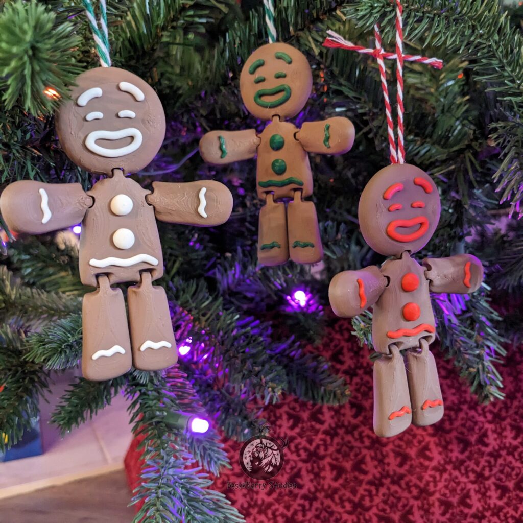 Trio of 3d printed gingerbread men ornaments hanging from a Christmas tree. All are brown, one with white icing, one with green, and one with red. Printed by BosonBerry Studios.